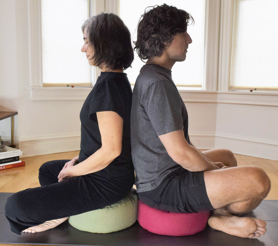 Man and woman sitting back-to-back on a zafu meditation cushion and yoga mat, each using a green and a red Zafu meditation pillow respectively.